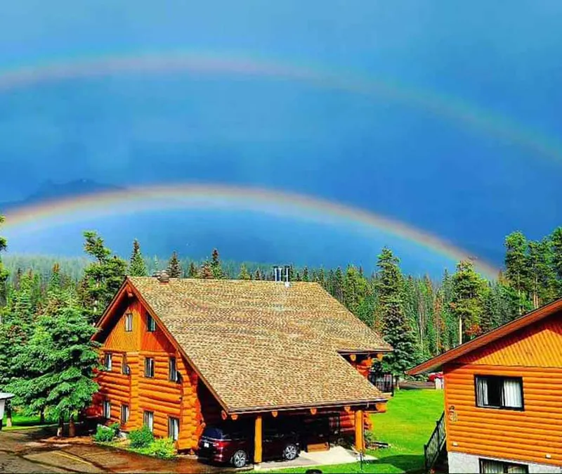 Log cabin chalets at Becker's Chalets with conifer forest and rainbow over blue alpine lake.