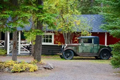 Log cabin with red walls and white trim beside weathered vintage truck amid tall pines at Johnston Canyon Lodge.