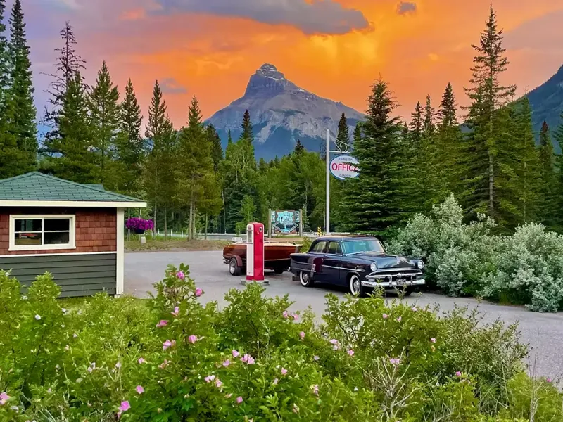 Johnston Canyon Lodge cabin with mountain peak, evergreens, and vintage truck at sunset.