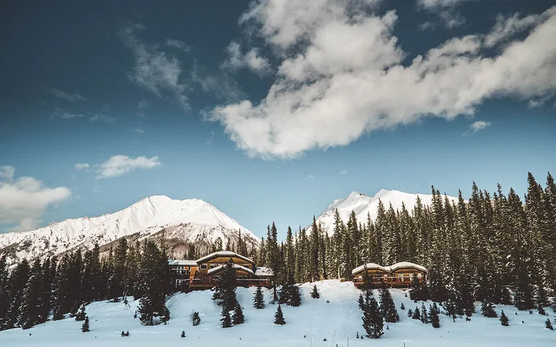 Mount Engadine Lodge's curved timber structures sit on a snowy hillside with snow-capped peaks and conifer forest behind.