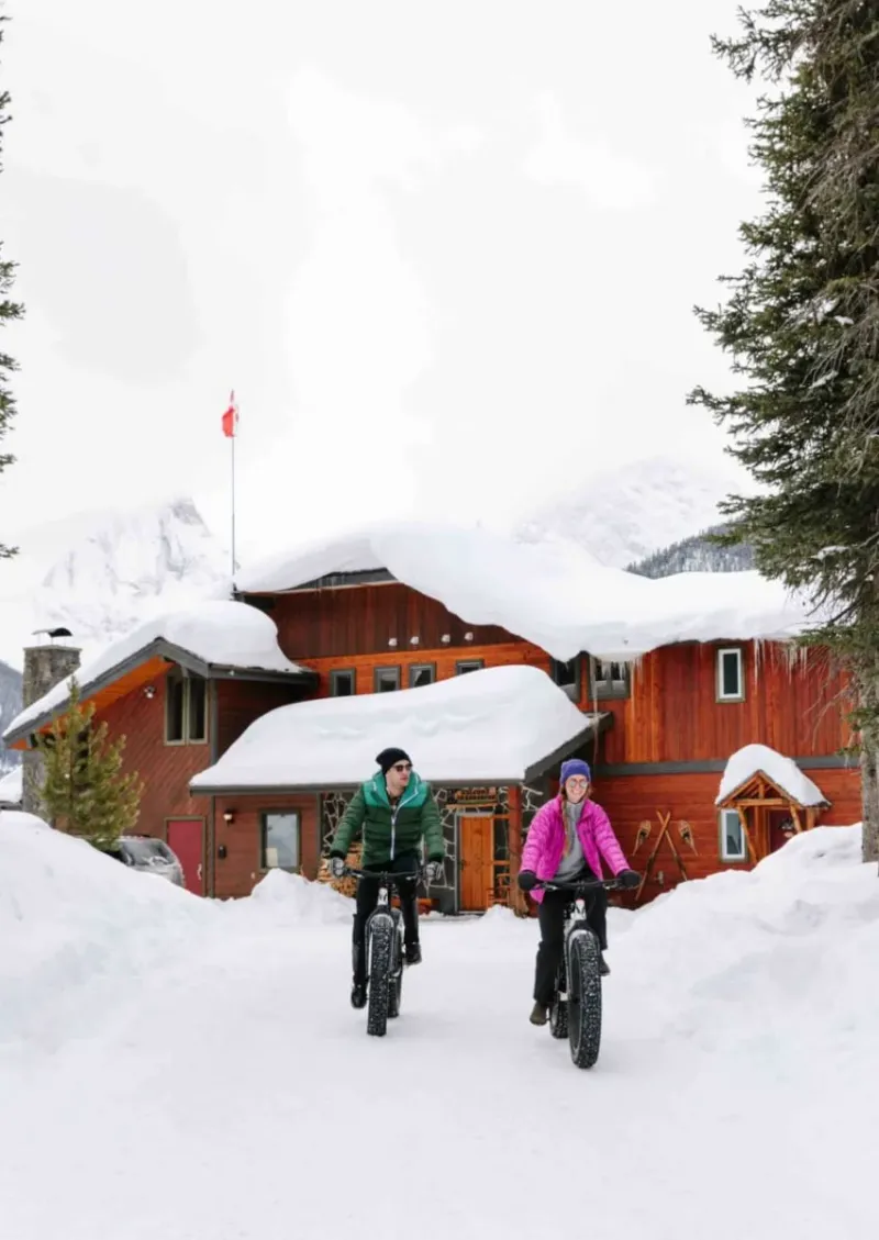 Two people riding fat bikes in snow in front of Mount Engadine Lodge's red wooden structure.