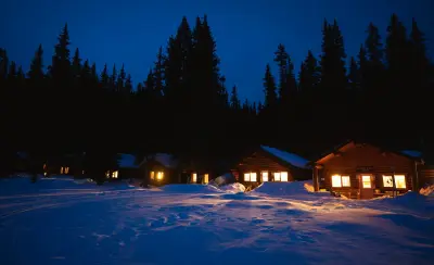 Log cabins at Shadow Lake Lodge glow warmly at dusk against deep snow and forested peaks in winter.