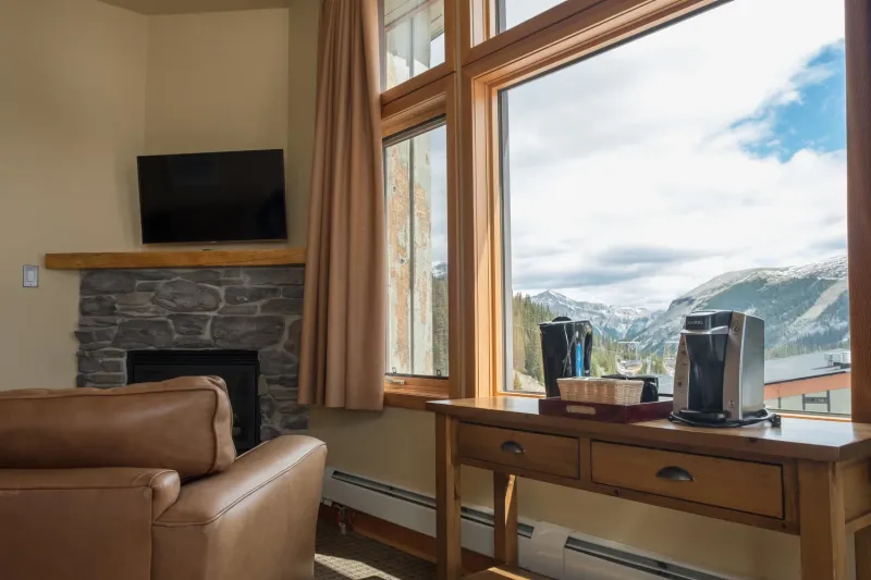 Sunshine Mountain Lodge suite with leather armchair, stone fireplace, and large windows overlooking snow-capped mountains.